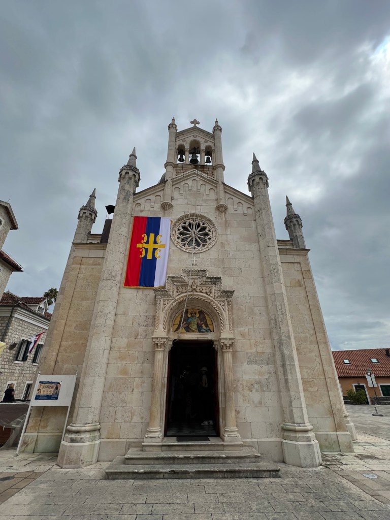 A photo of the front of a tiny old church with the door, a circular floral design window and the bell tower with 3 bells visible. 