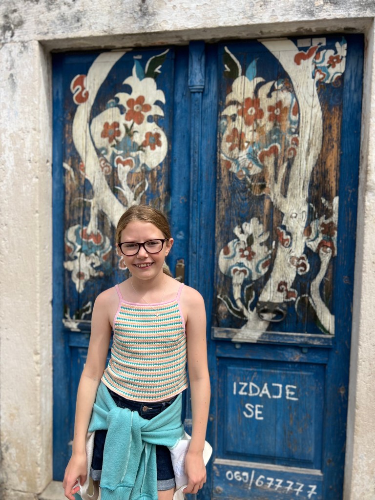 A 10 year old girl in a rainbow striped vest top, blue shorts and with a green jumper tied around her waist stood in front of an old wooden door painted blue with a floral design. 