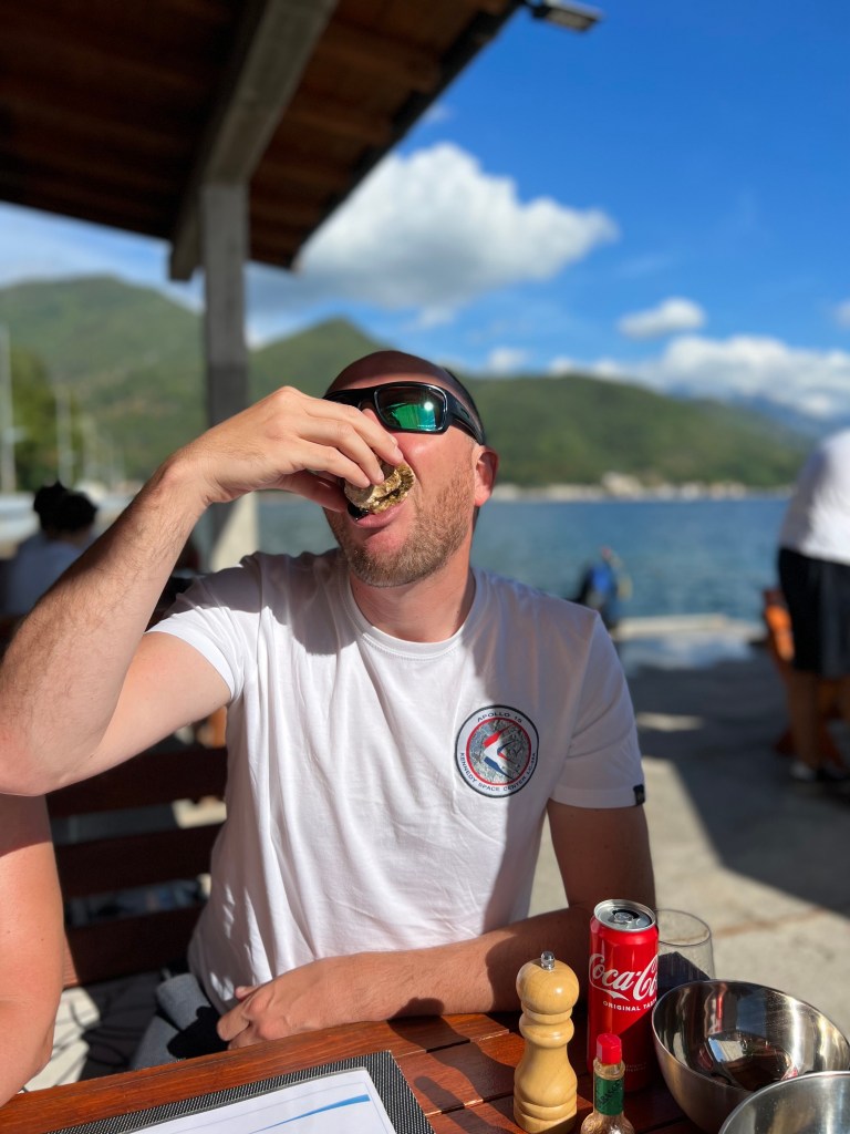 A man in a white t-shirt and sunglasses eating an oyster. 