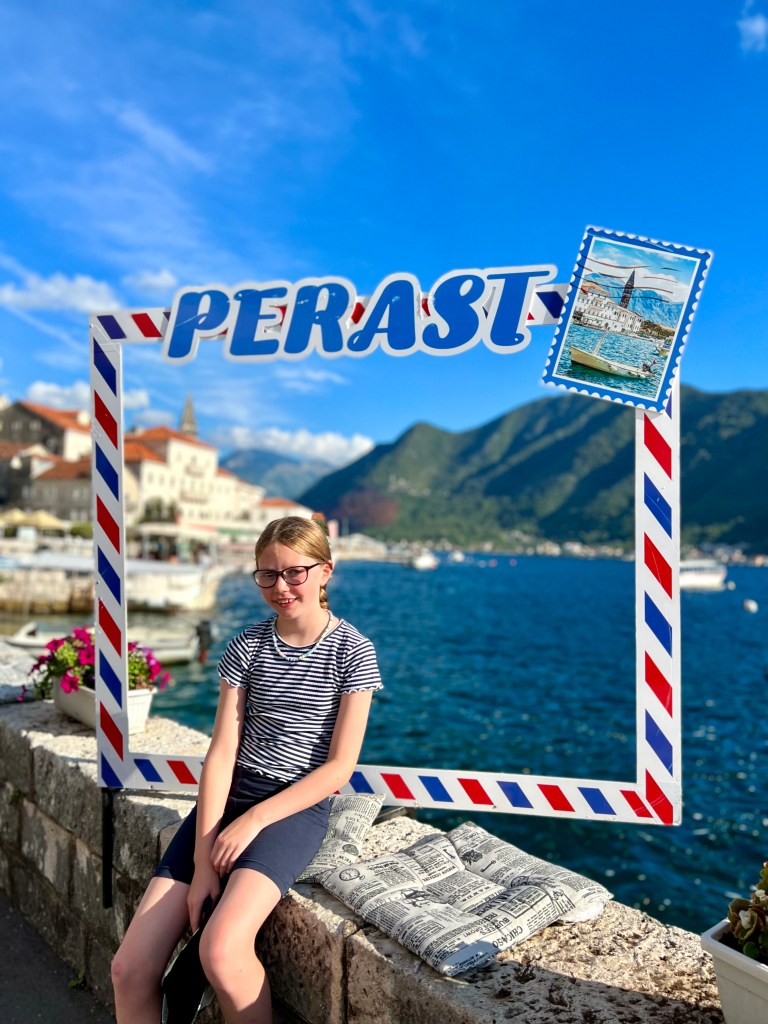 A 10 year old girl dressed in a blue and white striped t-shirt and blue shorts is sat on a wall in front of the Perast sign and has the sea and some of the town in the distance behind her. the persist sign is a red and blue striped frame that looks like an airmail envelope with a stamp in the top right corner and the word Perast at the top. 