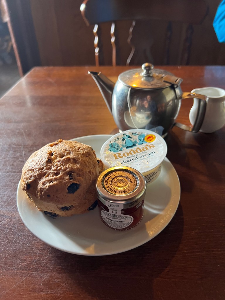 A plate containing a fruit scone, a small tub of Rodda's clotted cream and a small jar of jam. Behind the plate is a small silver teapot and a small white milk jug. 