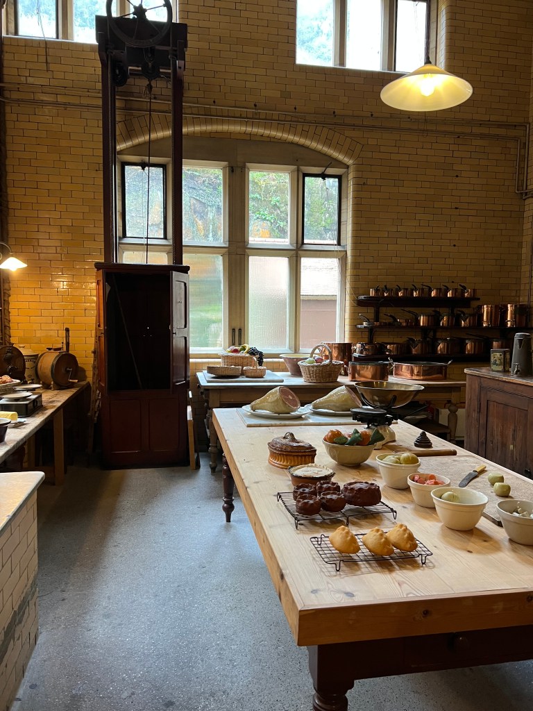 A photo of the large kitchen table with bowls of fake food. The far side of the room has large windows and in the left corner there is the dumb waiter with the pulley system above it. 