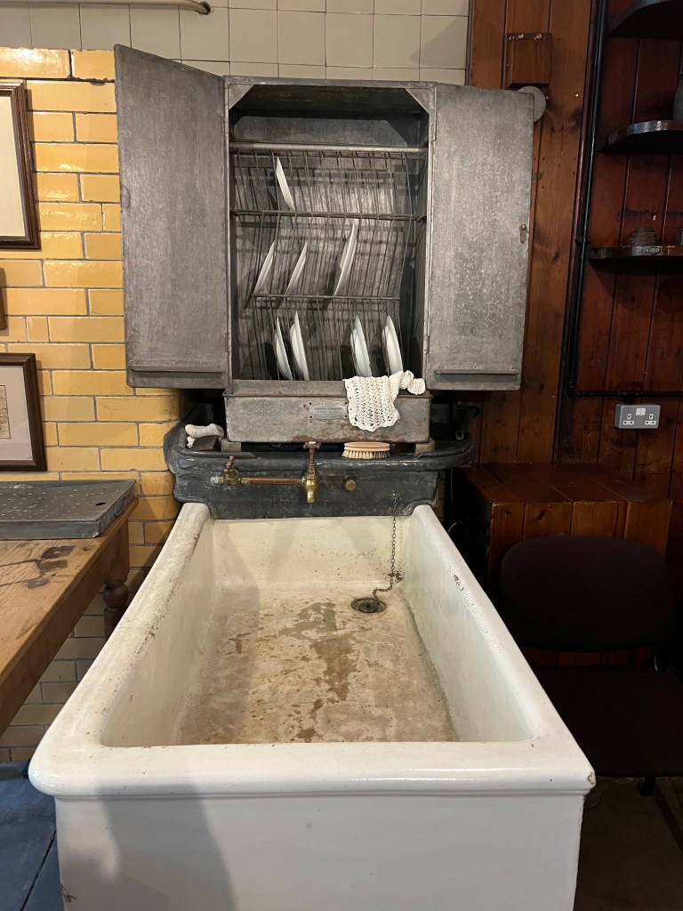 A large white Belfast sink with the dishwasher, a silver cupboard above it which is open showing plates on racks