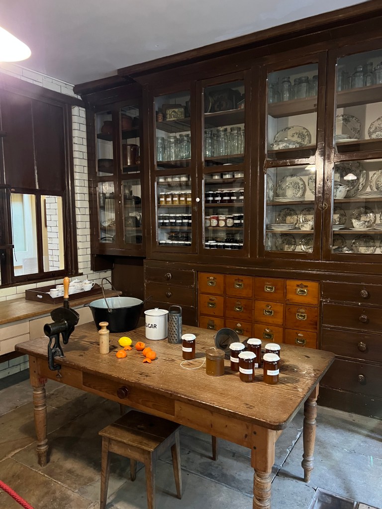A large wooden table with jars of marmalade and marmalade making equipment. behind are large glass fronted cupboards with crockery and jars of preserves. 