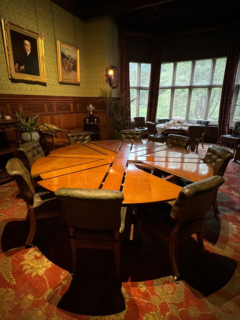 A large round wooden dining table with 8 green leather seats around it. The table is separated in to segments which is slightly open showing how the table can be expended to make it bigger. 