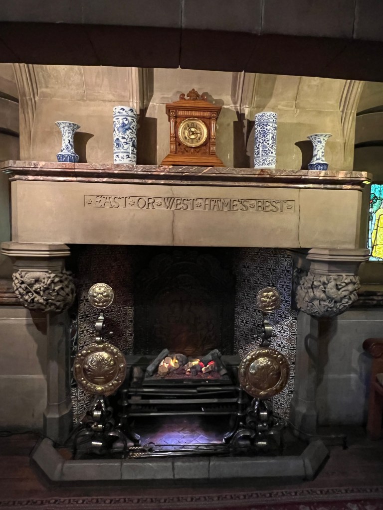 A large ornate stone fireplace. The mantle is inscribed with 'east or west hames best'. On top of the fireplace there are 4 blue and white vases and a carriage clock. 