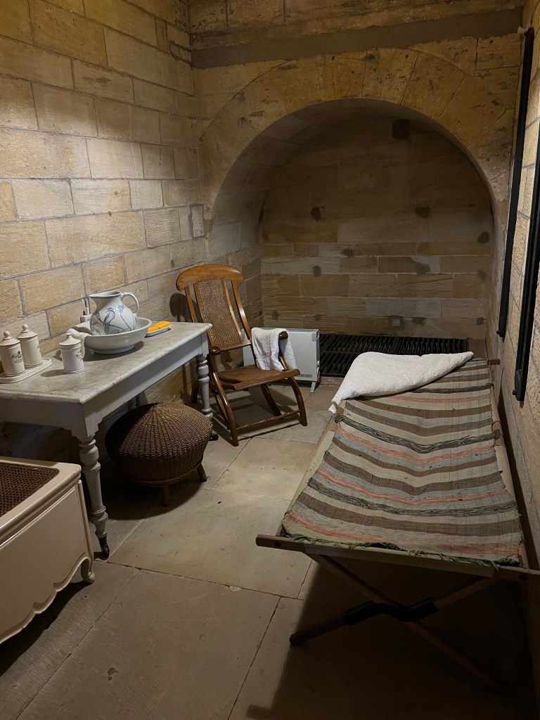 A stone room with a large stone archway on the far wall. There is a camp bed with a striped blanket on the right of the rooms and a marble table and wooden chair on the left. 