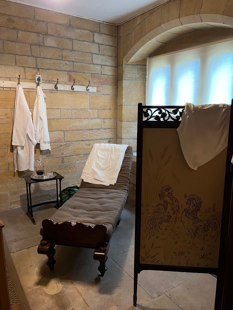 A stone room with a wooden sun lounger with a grey cushion. White bath robes are hung on the wall behind the lounger. 