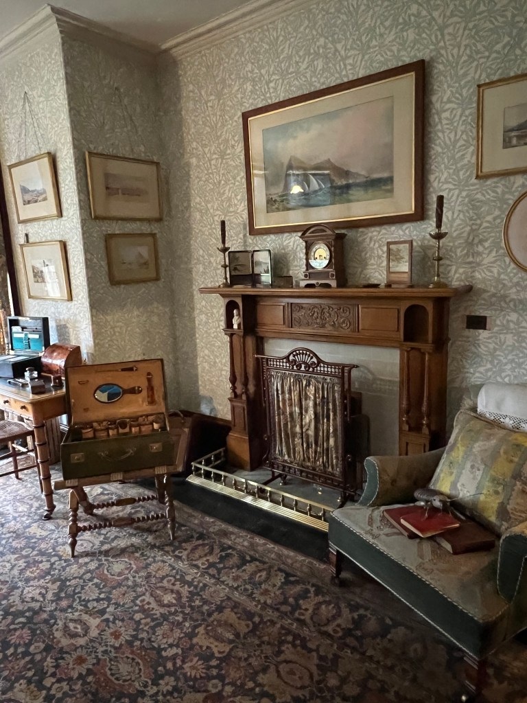 A photo of a wooden carved fireplace with green armchair to the left and an open wooden travelling trunk to the left. The wallpaper is green floral and the carpet is a blue and pink floral design. 