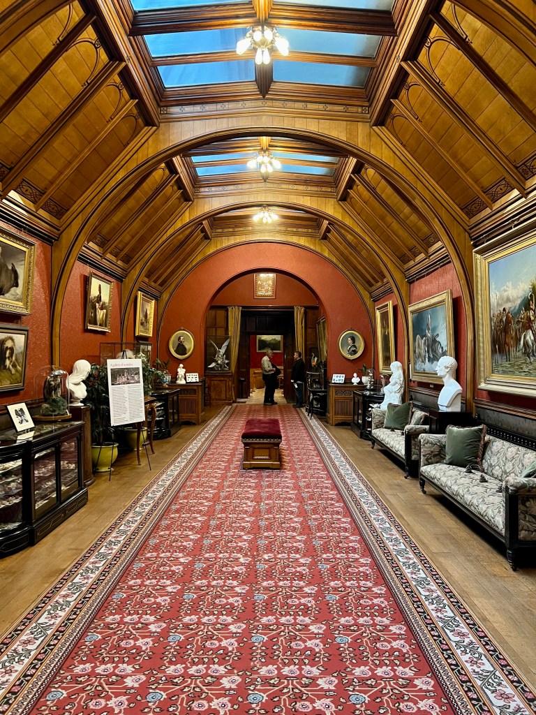 A photo of a gallery room with a wooden barrelled ceiling. The walls are painted terracotta and there is a terracotta floral runner on the floor. The walls are lined with large oil painting in gold frames. 