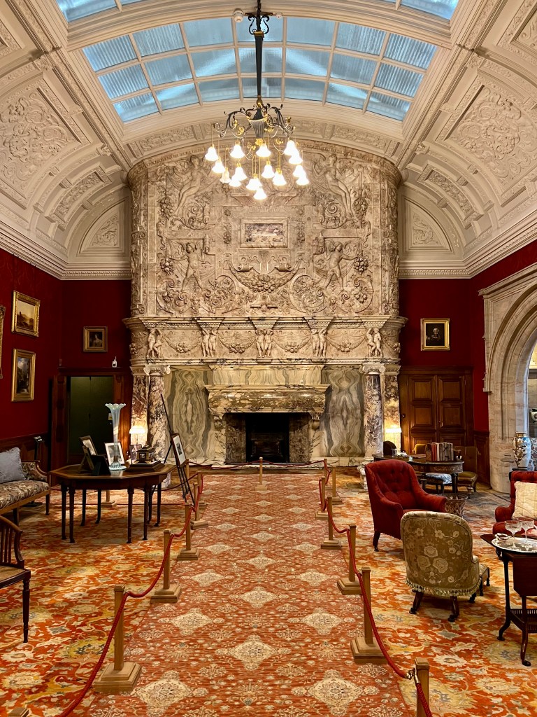 A large room with a domed ceiling  with  glass panels.  At the far end of the room is a huge, carved marble fireplace. The walls either side of the fireplace are painted dark red and the carpet is a red patterned design. There are various chairs and tables in the room in front of the fireplace. 