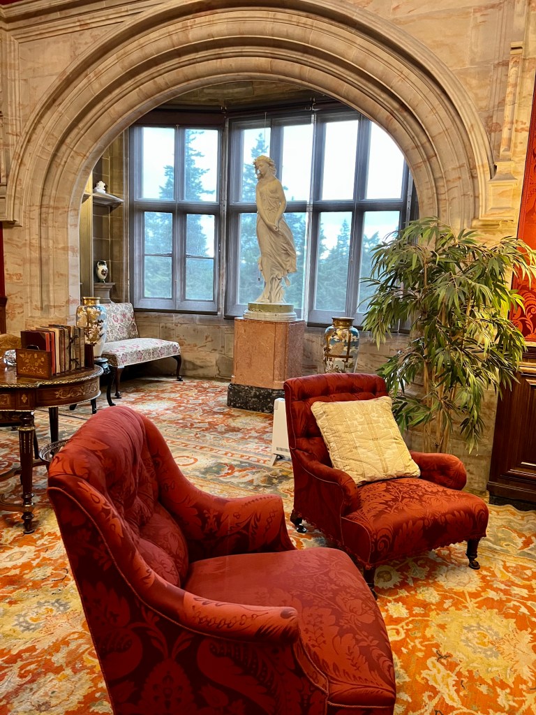 A photo of a large stone archway with a window behind it. There is a large marble statue on a plinth in the middle of the window alcove. In the foreground there are 2 red armchairs. 