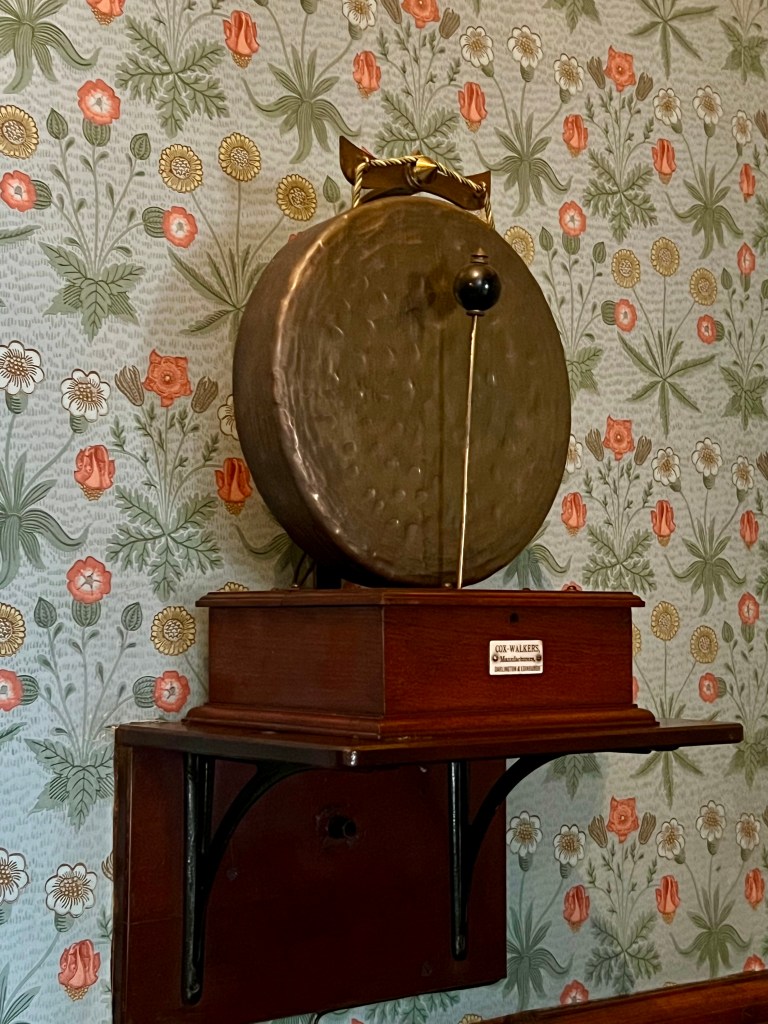 An automated brass dinner gong on the wall with green and red flower wallpaper.