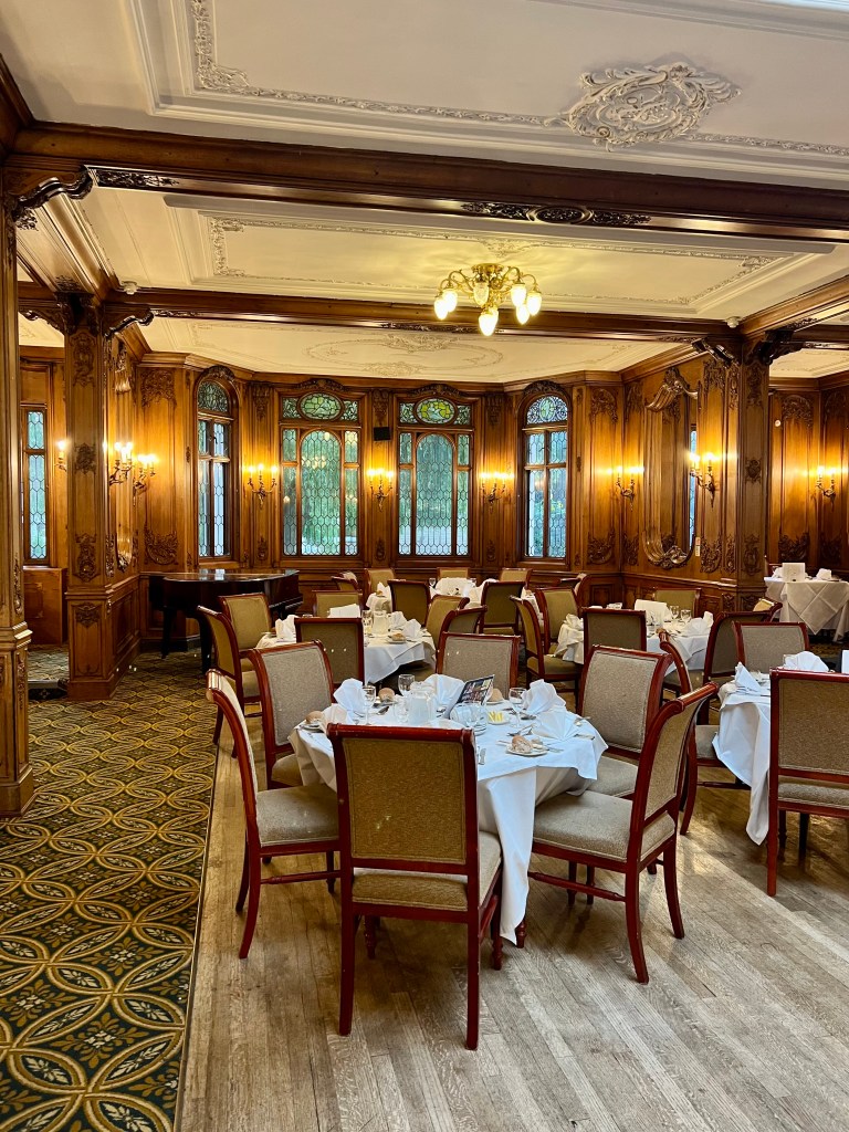 A shot of the Olympic dining room with several round tables set for dinner with white table cloths. The interior of the room is wood panelled with decorative windows.