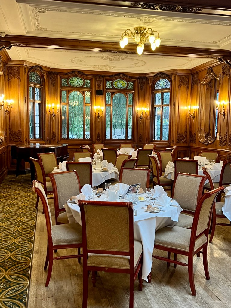 A shot of 4 round tables set for dinner for 6 with white table cloths. The tables sit in front of the ornate wooden panelling and windows of the Olympic dining room.