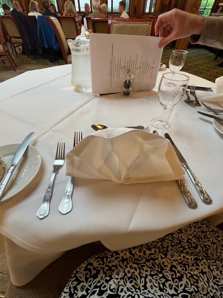 A round table with a white table cloth, table setting with 2 forks, 2 knives, a side plate with bread roll.