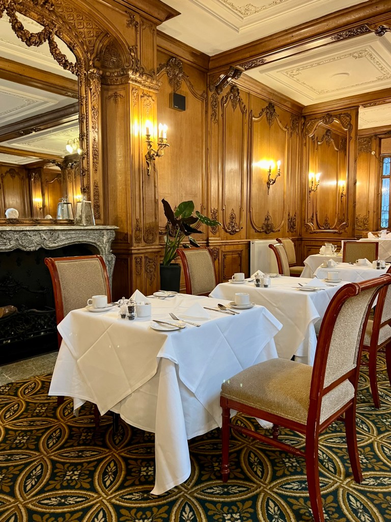 3 small square tables set for 2 for dinner with white table cloths. The front table is in front an ornate marble fireplace with a mirror above it.