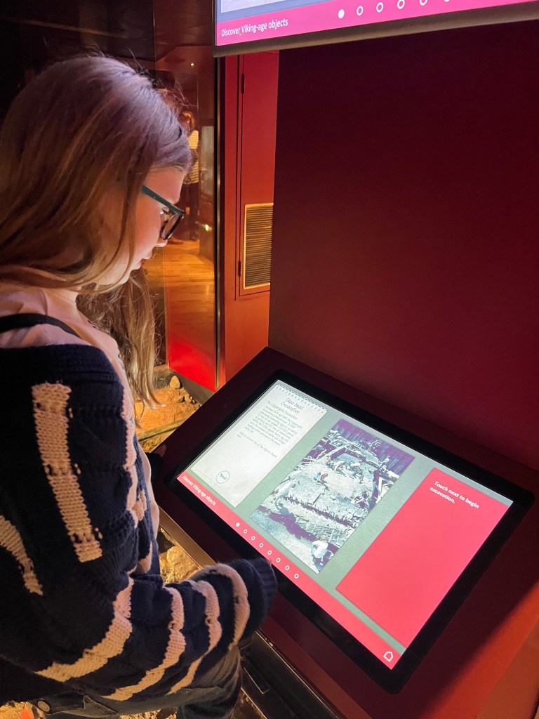 Pip, an 11 year old girl wearing a blue and white striped cardigan. She has long dark blonde hair and glasses and is looking at a computer screen.