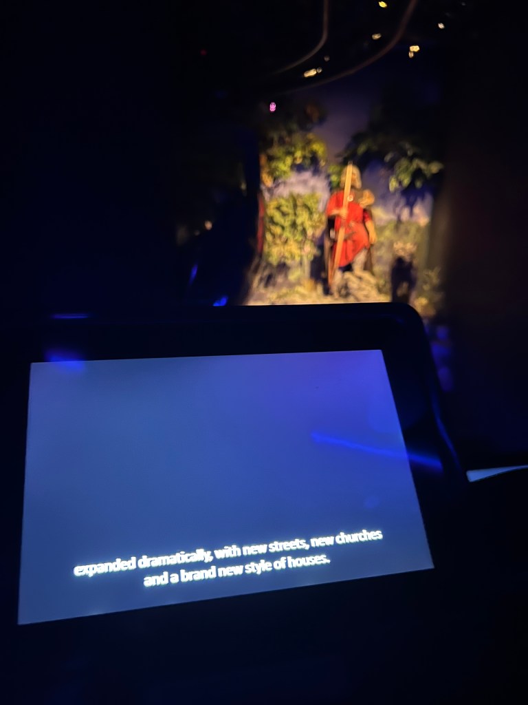 The screen on the car that takes you around the recreation of Jorvik York. On the screen there are the subtitles of what is being said slightly out of focus, in the background is a manikin of a viking man wearing red. 