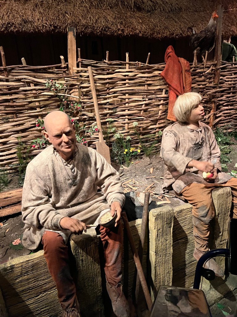 Two mannequins of Viking men sat on a wooden retaining wall that makes up the basement area of a house being built. The man on the left is bald and is wearing a beige robe and brown trousers, the man on the right is wearing similar clothes and has longer blonde hair. 