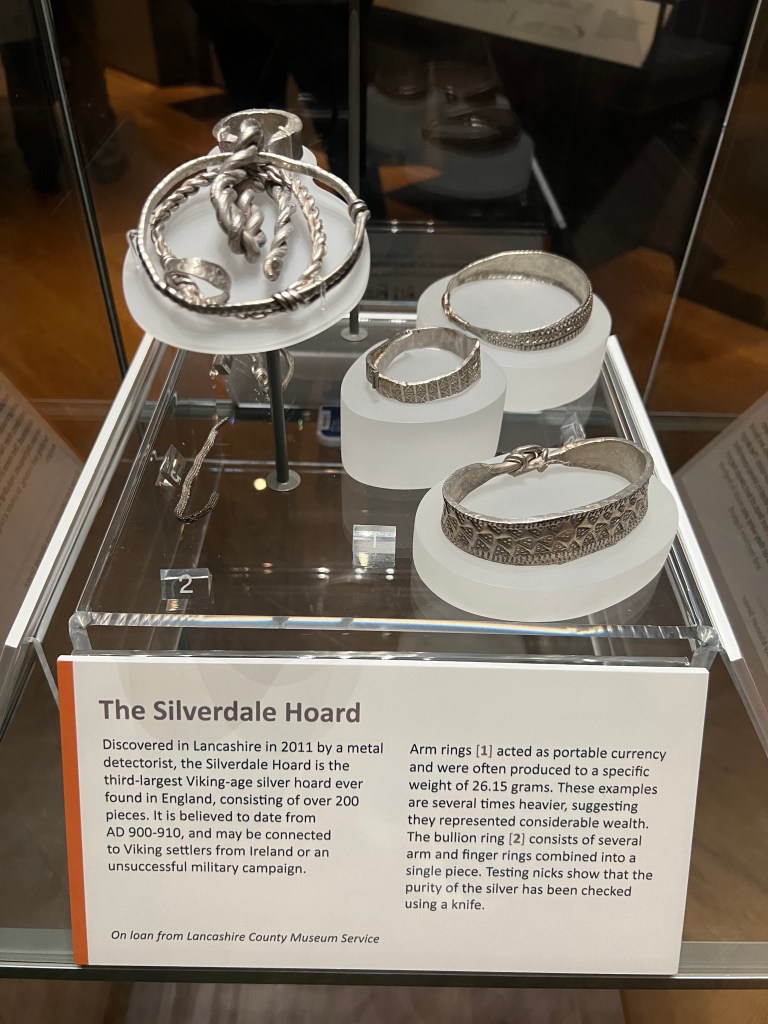 Various silver bracelets on display in a glass case. The information sign is titled The Silverdale Hoard and has text underneath which is too small to read. 