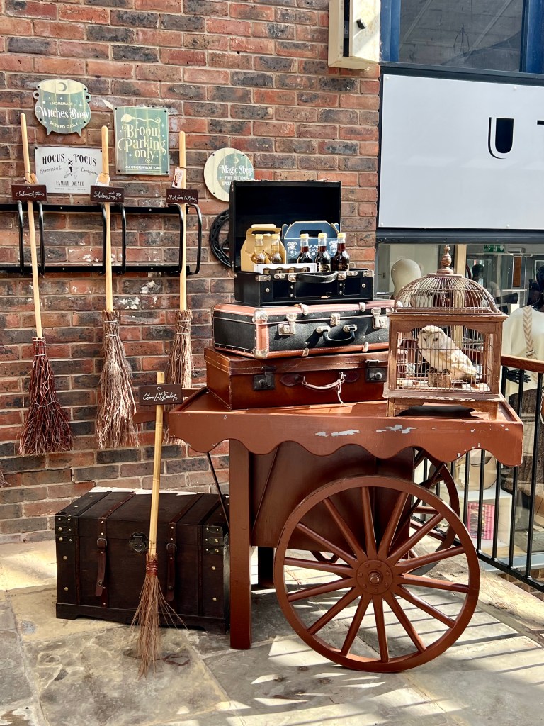 An old fashioned cart with old suitcases and a barn owl in a cage on top. Next to the cart there is a large wooden trunk and  a broomstick. Broomsticks are hanging on the brick wall behind the cart.