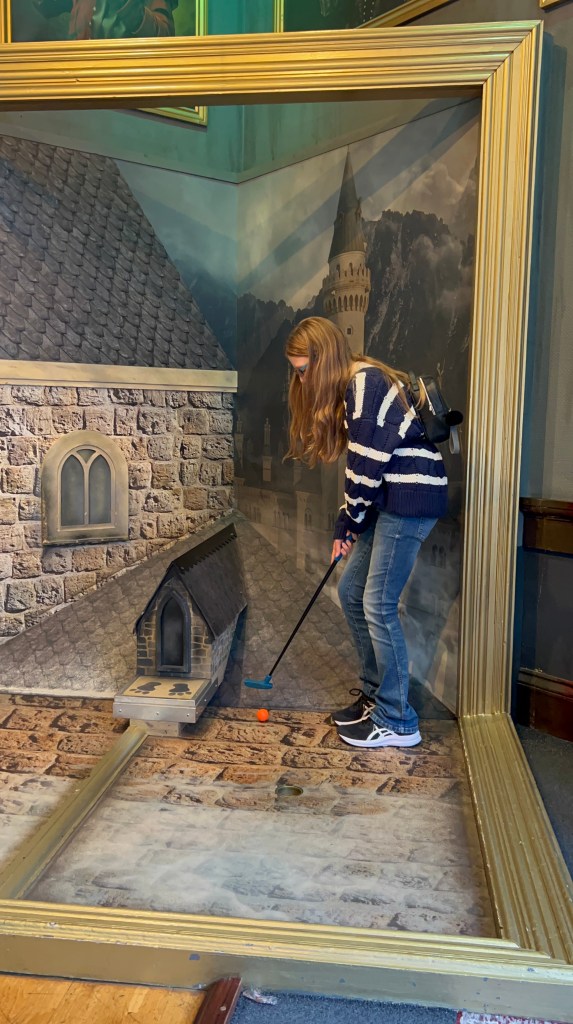 An 11 year old girl wearing blue jeans and a navy and white striped cardigan and a small black backpack is taking a golf shot on what looks like a large picture frame with a picture of the roof of a castle. 