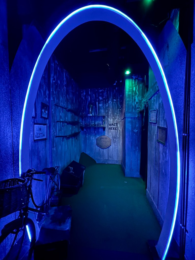 A blue lit room with wooden panelled walls. In the foreground is a blue neon arch way and an old bicycle. The room has broomsticks hanging on the walls. 