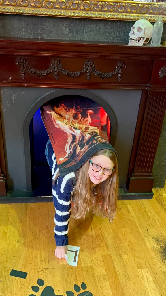 An 11 year old girl wearing blue jeans and a navy and white striped cardigan is crawling out of a secret exit in a  fireplace.