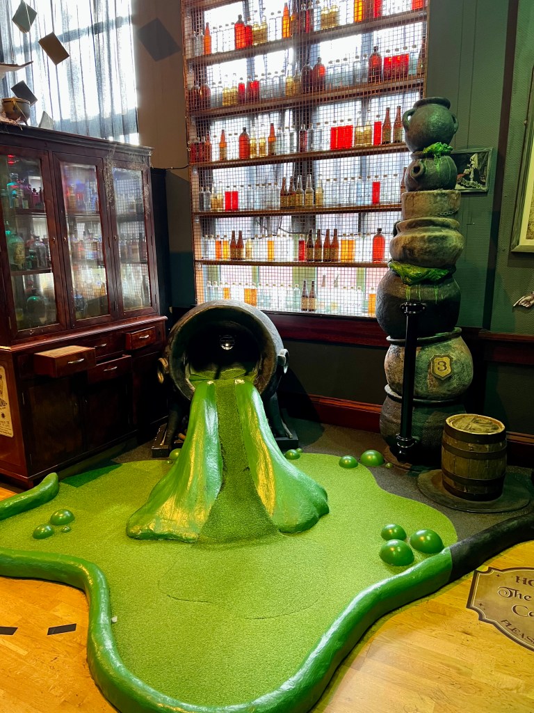 The photo shows on of the holes which is a large cauldron on it's side with green potion spilling and all over the floor. The hole is inside the cauldron. Behind the cauldron is a window with shelves of potion bottles in front. 
