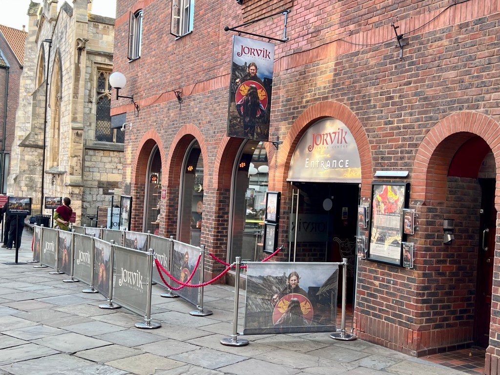 The entrance to the JORVIK Viking centre with 5 brick archways in a red brick building. The second archway is the entrance and has a large sign over it that says JORVIK Entrance. There is a banner hanging above the entrance with the word JORVIK and a picture of a viking man holding a red circular shield. 
In from the the building are barriers to direct the queues. 