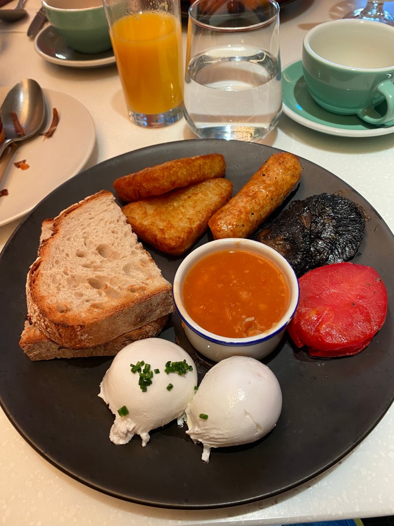 The photo shows a black plate with (clockwise from the top) hash browns, sausage, mushroom, tomato, two poached eggs and bread. There is a small ramekin of beans in the centre of the plate. In the back ground there is a glass of water, a green cup and saucer and a glass of orange juice.
