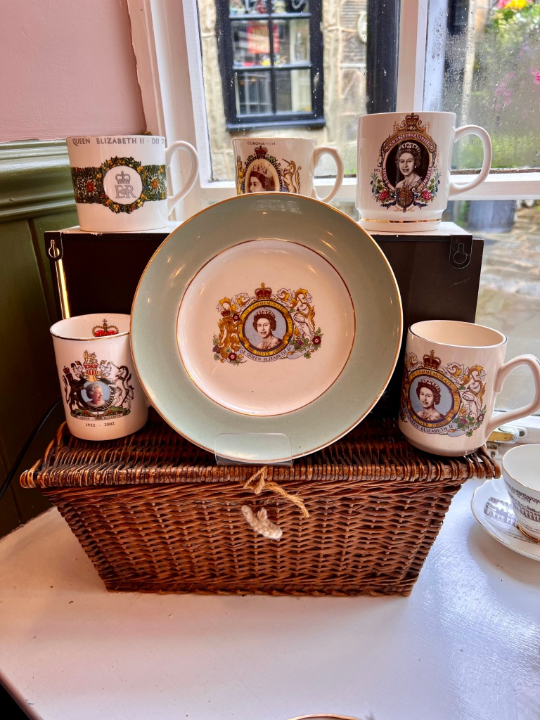 The photo shows a small wicker hamper. On top of it is displayed a selection of 5 different commemorative mugs with photos of the Queen on. In the centre of the display is a commemorative plate with a portrait of the Queen on and a green rim. 