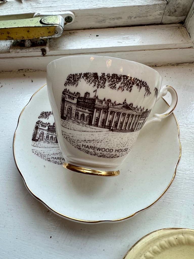 The photo shows a vintage cup lying on a saucer. The design on the cup is a black and white pen drawing of Harewood House. 