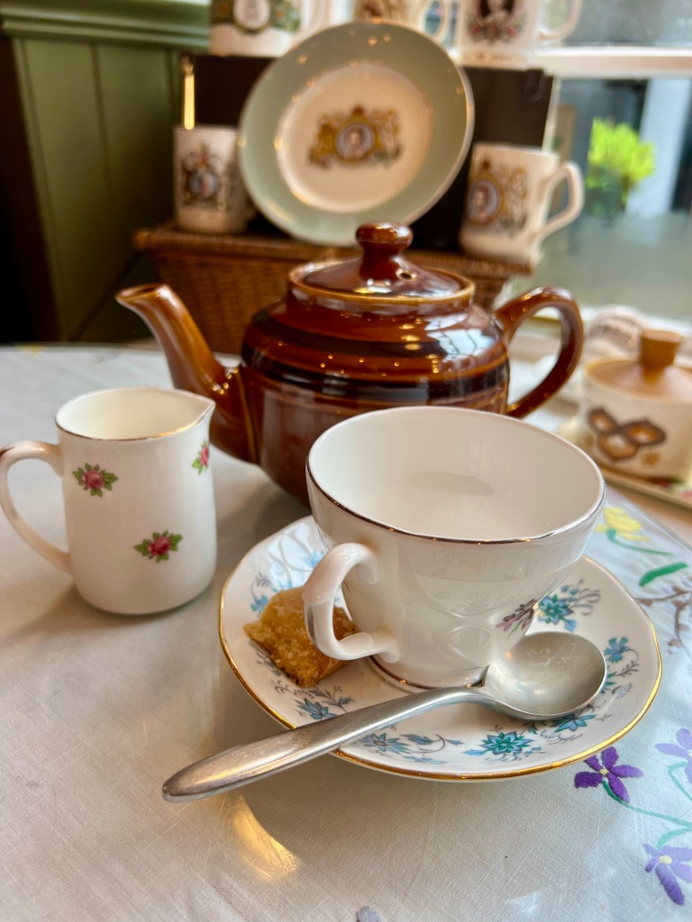 The photo shows a small brown teapot. in front is a white cup on a blue floral saucer and to the left is a small white milk jug with pink roses on it. 
