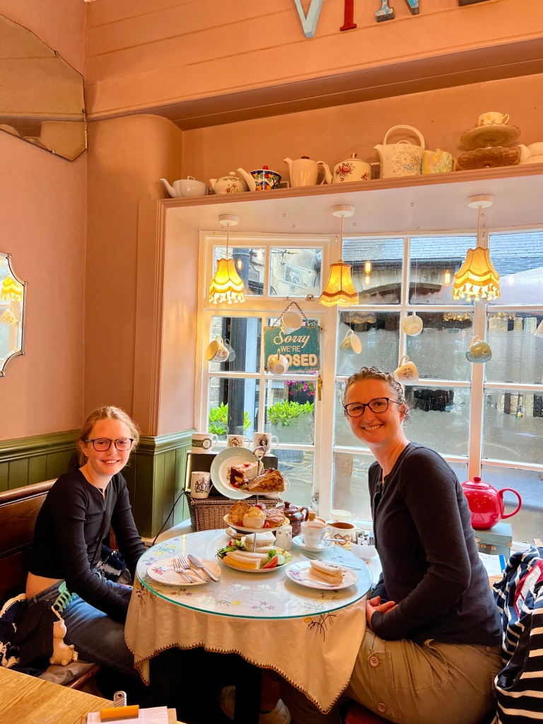 The photo shows Fee (on the right), a woman in her forties wearing khaki trousers and a black long sleeved top. She has brown hair tied back and glasses. On the left is Pippa, an 11 year old girl wearing blue jeans, a long sleeved black top, she has dark blonde hair tied back and glasses. 
They are sat at a table for 2 with a white table cloth. The table has a cake stand with sandwiches and cakes on it. They are sat in front of a bay window. 