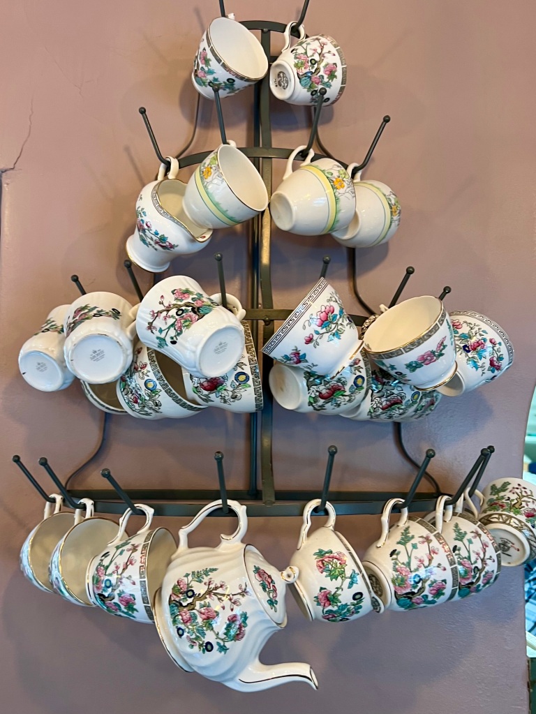 The photo shows an assortment of vintage cups with a variety of floral designs hanging from a tree shaped rack. A floral teapot hangs from the bottom centre. 