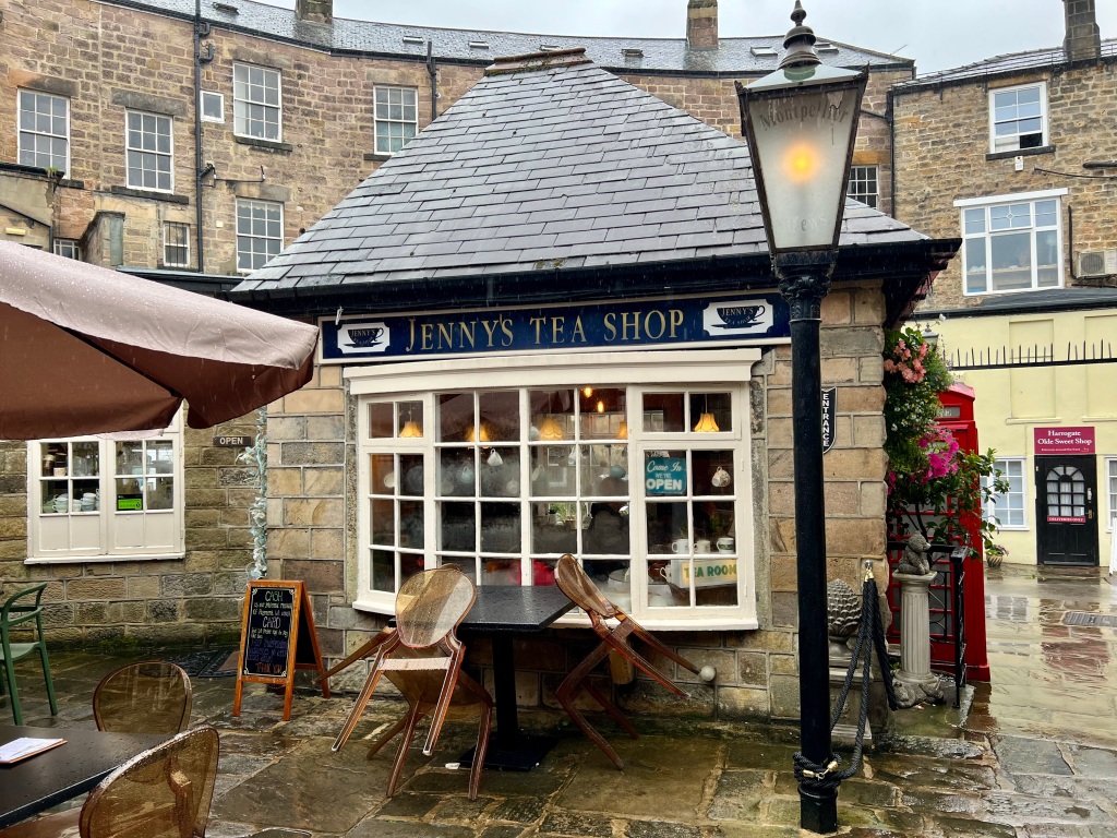The photo shows Jenny's Tea Shop, a small, one storey stone building with a black slate roof and bay window. Above the window there is a blue sign  which says Jenny's Tea Shop.
Behind the Tea shop are other larger stone buildings and in front is an old fashioned gas street lamp, some tables and chairs and part of a table umbrella. To the right of the tea shop is a red phone box. 