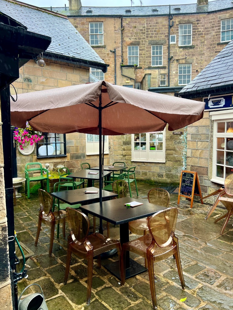 The photo shows the courtyard outside of Jenny's Tea Shop with stone paving flagstones and surrounded by stone buildings. The table at the front of the photo has a large pink umbrella. 