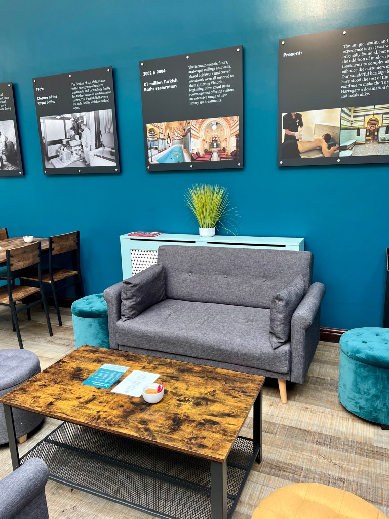 A grey sofa in the reception area with a wooden coffee table in front and two turquoise round stools on either side. The wall behind is painted dark turquoise and there is a row of signs on the wall showing old photos and information about the history of the baths.