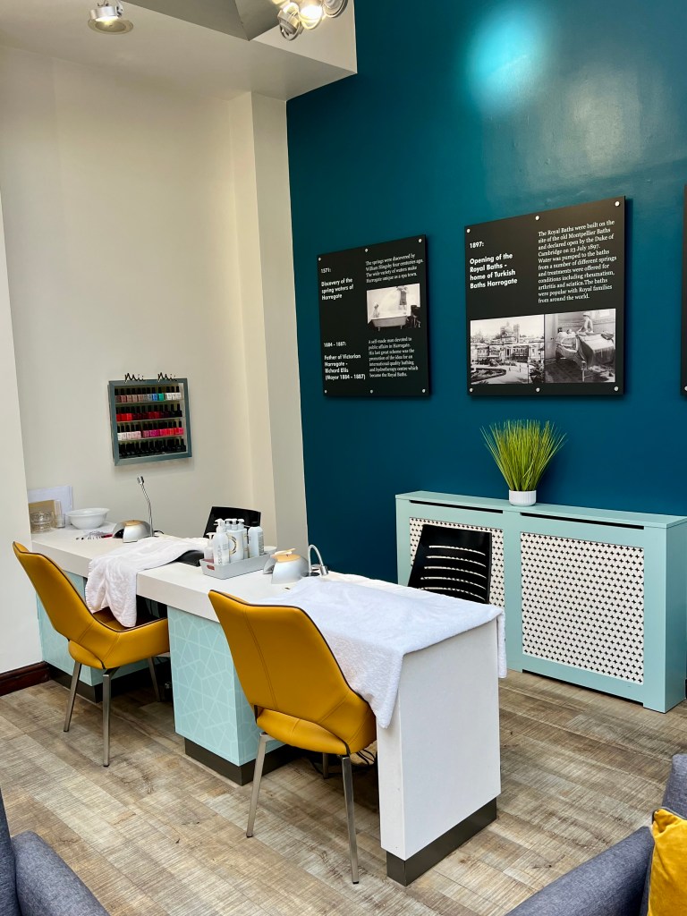 The two nail stations in the reception area of the Turkish Baths. They both have a yellow chair sat at them and are covered in white towels and various equipment. The wall behind is painted a dark blue and there are two large signs on the wall with old photos and information about the baths.