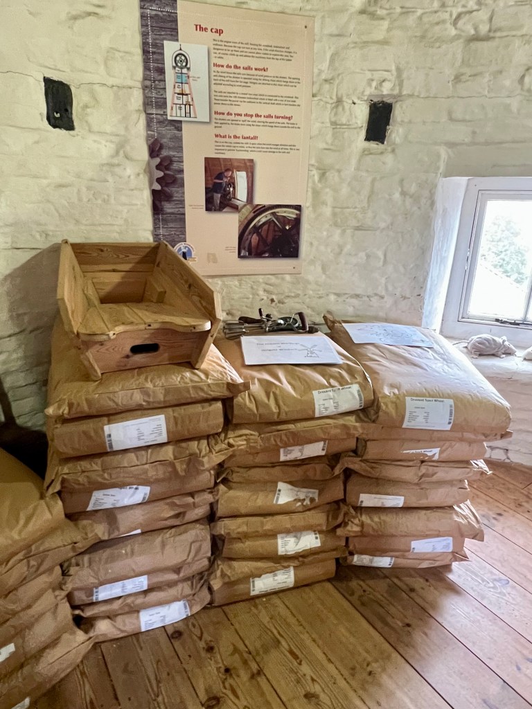 The photo shows big brown bags of grain stacked on a wooden floor in front of a white stone wall. On the wall is a large information sign with details about the windmill and there is a small window to the right of the photo.