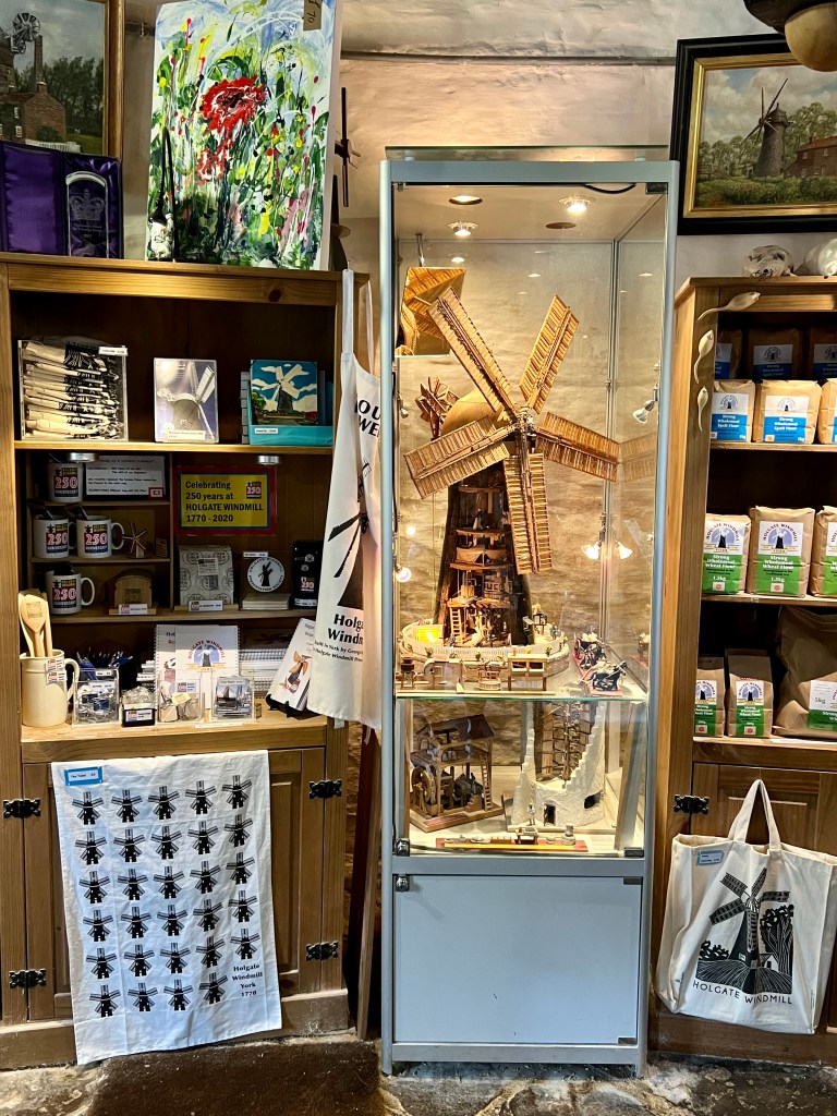 The photo shows the gift shop. In the centre there is a thin glass cabinet which contains a model of a windmill without the front wall so you can see inside.
To the right are wooden shelves containing brown paper bags of flour. To the left are wooden shelves containing souvenirs such as mugs and magnets. A tea towel and tote bag are hanging from the shelves. Above the left shelves is a large canvas painting of a poppy.