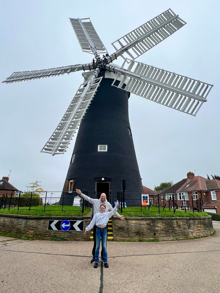 The photo shows Holgate windmill. The stone body of the windmill is painted black and it has 5 white sails. The windmill is stood on a road about in the middle of the road, behind it is some red brick semi-detached houses.
In front of the windmill in the middle of the road stands Andy; a tall man in his forties wearing black trousers, white trainers and a light grey hoodie. In front of him is Pip; an 11 year old girl wearing blue jeans, black shoes and a grey sweatshirt. She has light brown hair tied back and glasses. They are stood with their arms out to the side like windmill sails.