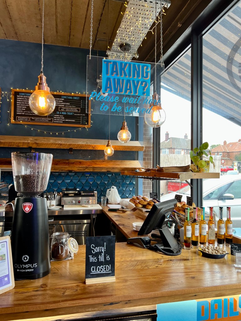 The wooden counter at Bluebird Bakery and Cafe. On the counter is a small blackboard sign says 'sorry this till is closed'. There is also coffee beans in a grinding machine and various bottles and pasties. 