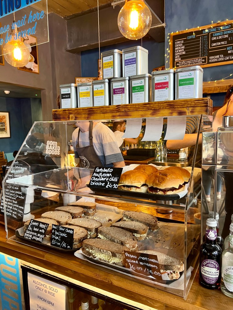 A glass display case on the counter containing and assortment of different sandwiches. There is a selection of teas in metal canisters stacked on top of the display case. 