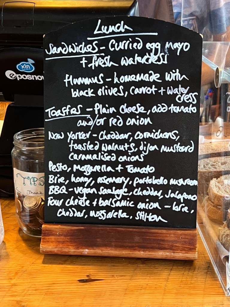 The lunch menu written on a small blackboard. 
Lunch
Sandwiches - Curried egg mayo and fresh watercress.
Hummus- homemade with black olives, carrot and watercress. 
Toasties - Plain cheese, add tomatoes and/or red onion.
New Yorker - Cheddar, cornichons, toasted walnuts, dijon mustard, caramelised onions
Peston, Mozzarella and tomato
Brie, honey, rosemary, portabello mushroom
BBQ- Vegan sausage, cheddar, jalapeño
Four cheese and balsamic onion - brie, cheddar, mozzarella and stilton
