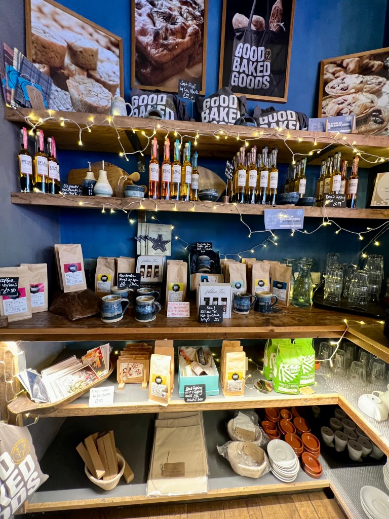 5 wooden shelves in front of a blue wall. The shelves are stocked with items that can be purchased from the bakery such as bottles of oil, flour and teas. 
