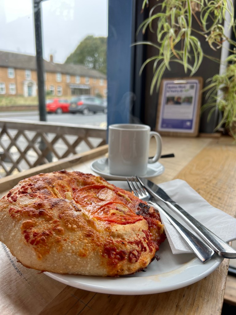 The photo shows a small flatbread topped with tomato and cheese on a small plate with a knife and fork. Behind the plate is a white mug. 