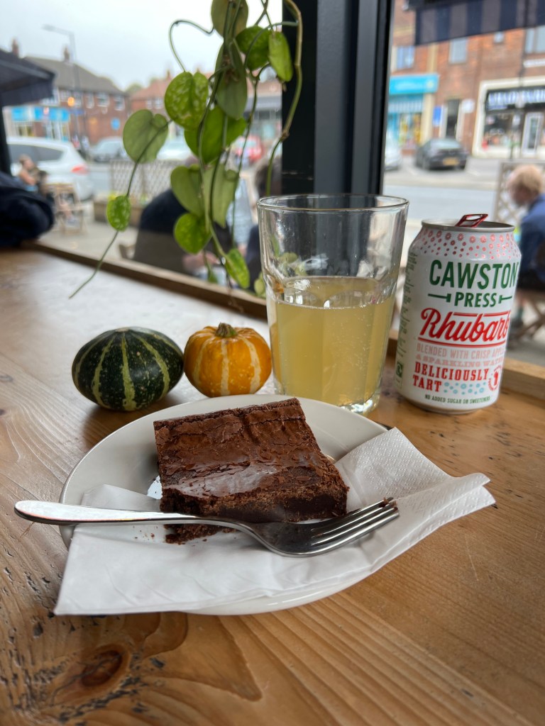 The photo shows a square brownie on a plate with a fork. Behind it is a can of Cawston press Rhubarb and a glass half full of the drink. 